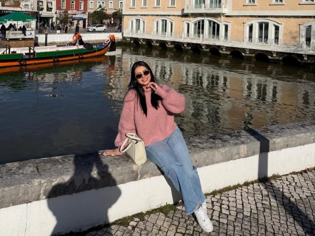 IsabellaJames with dark hair, pink sweater, blue jeans, white sneakers, holding beige bag, leaning on stone wall by canal, gondola in background, wearing sunglasses, smiling.