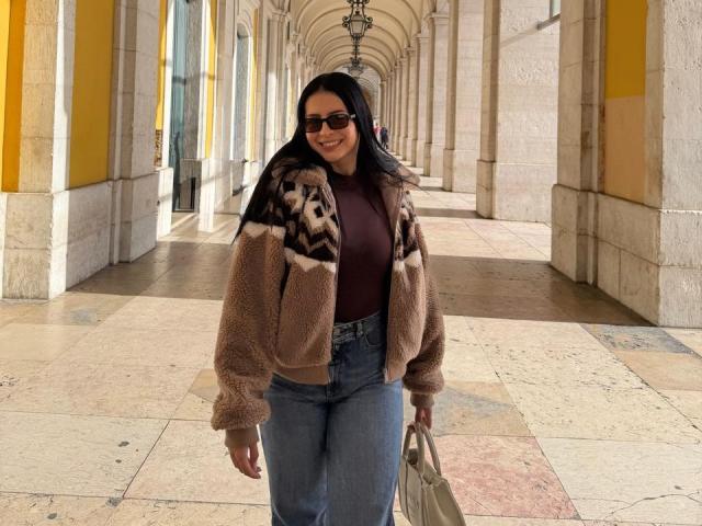 Isabella James, com cabelos longos e pretos, vestindo uma jaqueta marrom felpuda com estampa, uma camiseta escura, calças jeans azuis de cintura alta, segurando uma bolsa bege, sorrindo e usando óculos de sol escuros..