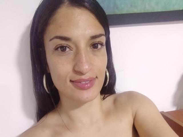 VeronicaDell has medium brown skin, long black hair, brown eyes, and pink lips. She's smiling, wearing hoop earrings, and has small, visible freckles on her nose.