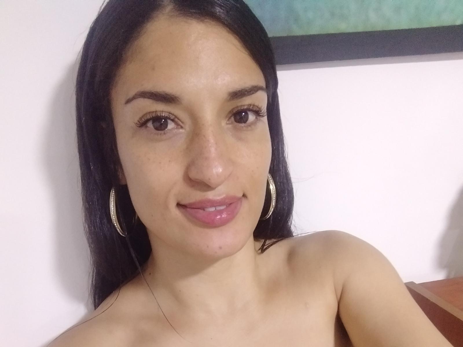Veronica Dell tem a pele marrom clara, cabelos pretos longos, olhos castanhos e lábios rosados. Ela está sorrindo, usando brincos grandes e tem pequenas sardas visíveis no nariz..