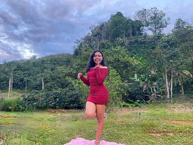 JuliaBennett with long black hair, smiling, wearing a tight red dress, standing on one leg, grassy field, pink cloth, green forest, cloudy sky, holding green leaf.