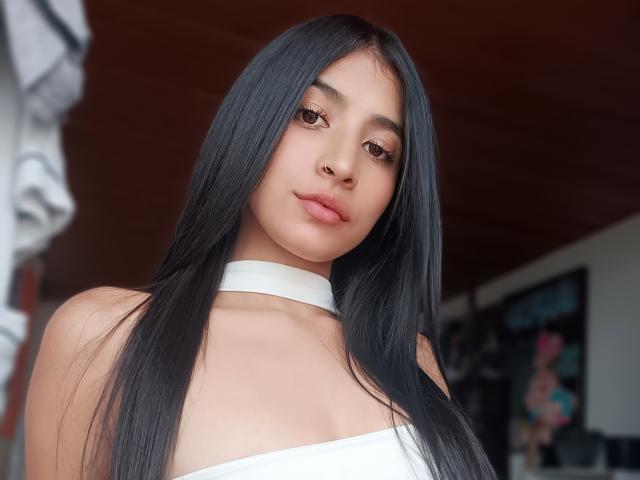 NaiaSanders with long black hair, white strapless top, and white choker, has full pink lips and brown eyes, looking at camera with a slight, confident smile.