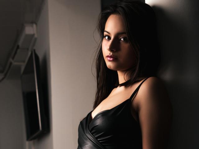 LunaBellee with dark brown hair, wearing a black leather dress, standing against a wall, looking at the camera with a slight, confident smile, showing her medium-sized tits.