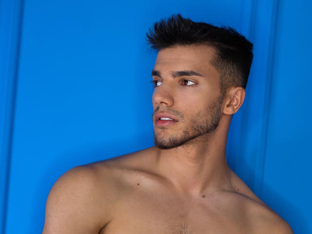 DominicValente, shirtless with a toned upper body, has short dark hair, a trimmed beard, and brown eyes, staring off to the side against a bright blue background.