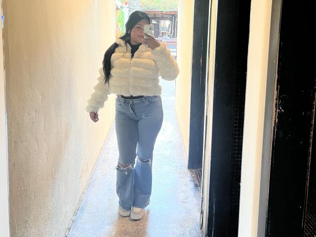 AbrilLuxurie met lang zwart haar, gekleed in een pluizige witte jas, versleten blauwe jeans en witte laarzen, maakt een spiegelzelfportret, waarbij haar achterwerk in de reflectie te zien is..