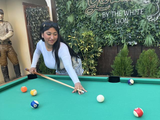 AbrilLuxurie, with long black hair, blue top, and jeans, leans over a green pool table, aiming a cue stick at balls, showing her tits slightly through her shirt.