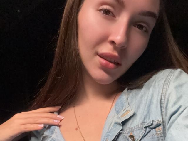 AquaLady has light skin, brown hair, and is wearing a denim jacket with a gold necklace. She's looking at the camera with slightly parted lips, holding her jacket.