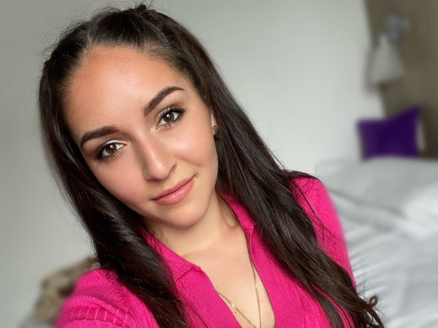 AquaLady with long dark hair, pink shirt, and subtle makeup, smiling at the camera. She's got cute brown eyes and a slight tan. Her tits are not visible.
