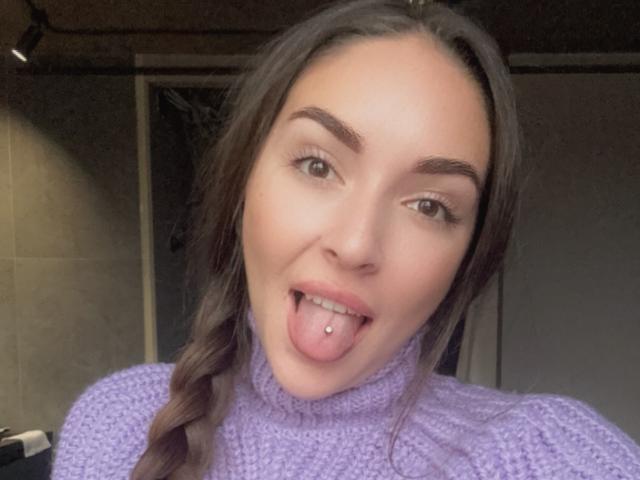 AquaLady with long brown hair, purple turtleneck, tongue out, showing a tongue piercing, green eyes, and a slight smile, in a selfie.