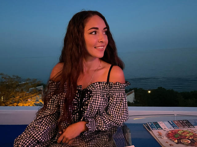 AquaLady with long brown hair, smiling, wearing an off-shoulder black and white checkered dress, sitting by a blue-topped table, dusk by the ocean. Her tits are subtle.