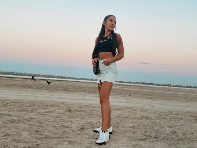 AquaLady with long brown hair, tan skin, small tits, wearing a black sports bra, white denim shorts, white sneakers, holding a camera, standing on a sandy beach at sunset.