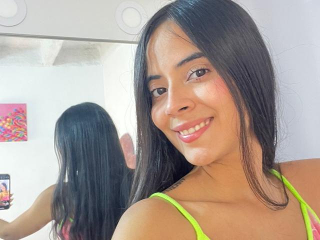 TamaraPeach with long black hair, smiling, wearing a neon green top, showing her shiny lips and smooth brown skin in a mirror selfie. Her tits are slightly visible.
