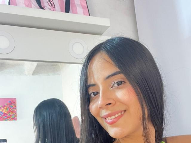 TamaraPeach with long black hair, brown eyes, and pink lipstick, smiling at the camera, reflection in the mirror, white shelf with pink bag, and colorful wall art in background.