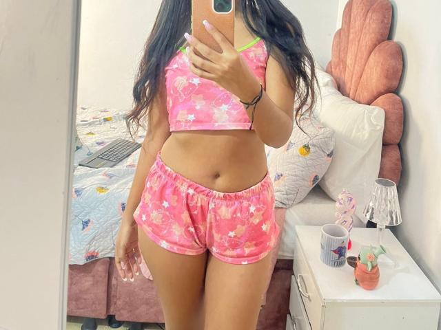 TamaraPeach in pink floral crop top and shorts, holding phone, showing tan skin, long brown hair, small green bra strap, pink headboard, white bed, and pink bedside table.