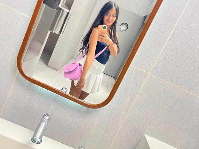 TamaraPeach with long black hair, smiling, holding phone, wearing black top, white skirt, pink purse, standing in bathroom, mirror reflection, showing her ass.
