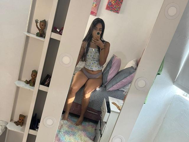 TamaraPeach stands in a mirror, taking a selfie with her phone. She has long, dark hair and wears a light-colored, floral-patterned top with gray panties. Her toned, tan-skinned legs are prominent. She's in a bedroom with a bed, white shelves, and colorful artwork. The room has a colorful rug and pink pillows. The mirror has circular marks. Light from a window is on the right. Slang: "TamaraPeach is looking real cute."