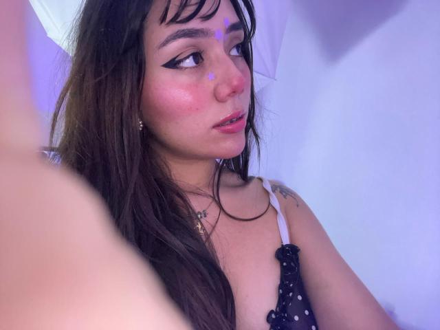 AlannaJonson with long brown hair, black eyeliner, pink blush, and a black polka dot top, looking to the side with a slight smile, showing small tattoos on her chest.