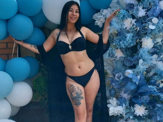SexyLubov with black bra and panties, long black hair, tattoos on thigh, holding blue and white balloons, standing in front of blue and white floral arrangement, smiling.