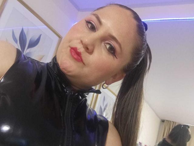 TiffanyStor con labios fruncidos, lápiz labial rojo y una blusa de látex negro brillante, mejillas abultadas, una coleta marrón, aros grandes y delineador de ojos llamativo, en un selfie..