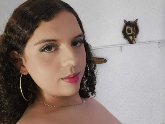 CatleyaNoire hat lockiges, braunes Haar, grüne Augen und pinken Lippenstift. Sie trägt Ohrringe im Hula-Hoop-Stil und eine Halskette, mit einem kleinen Eulen-Ornament an einer weißen Wand..