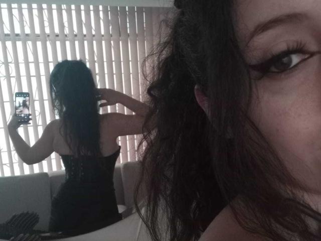 KendraSimons with wavy black hair, wearing a black lace corset, taking a selfie in a mirror showing her back, holding a phone, her ass visible in the reflection.