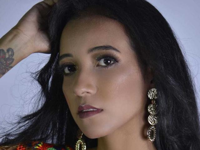 AshleyMillet has dark, wavy hair, brown eyes, and wears ornate gold earrings. She has a tattoo on her left arm and dark maroon lipstick.