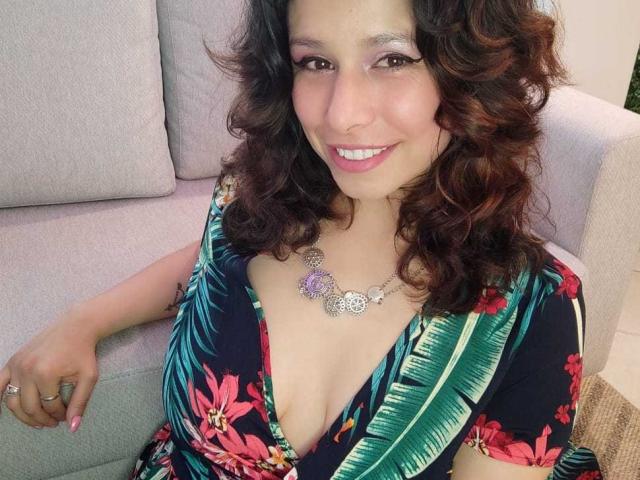SarahWelinton with curly brown hair, pink lipstick, and purple eye makeup, smiling, wearing a floral dress with a deep cleavage, showing her tits, and a purple necklace.