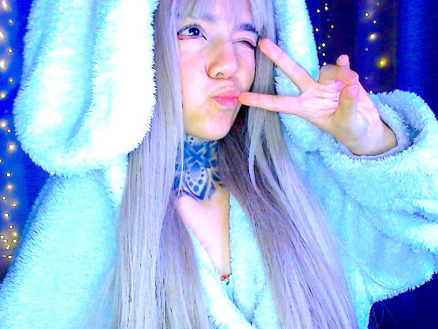 LannieGonzales with long gray hair, wearing a fluffy blue bunny hoodie, winking, and making a peace sign with her pink tongue out. Glowing blue light highlights her cute face.
