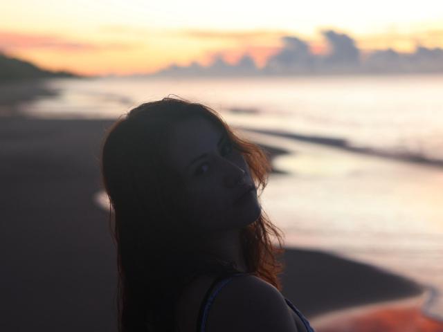 MalenaJones with dark, wavy hair looking back over her shoulder, soft sunset light, blurry beach background, wearing a blue tank top, subtle blush, serious expression.