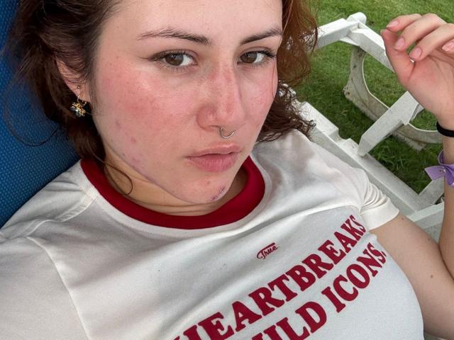 MalenaJones has pale skin, brown curly hair, a nose ring, and is wearing a white shirt with red text. She's lying down with a pouty expression and blue chair.