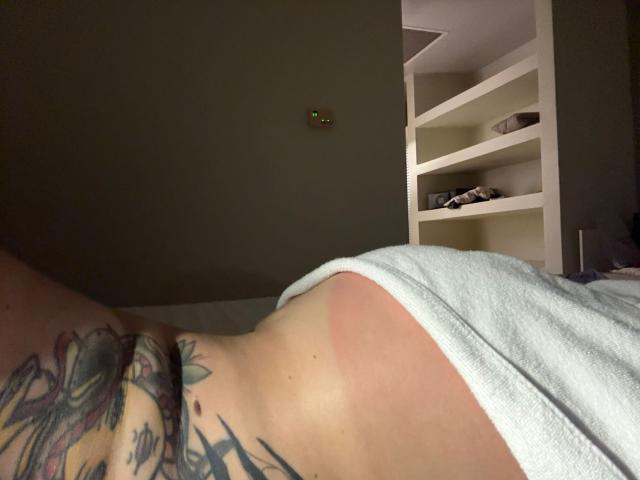 MalenaJones' lower back with a colorful tattoo, showing a red tan line, lying on a bed with a white towel, in a dimly lit room.
