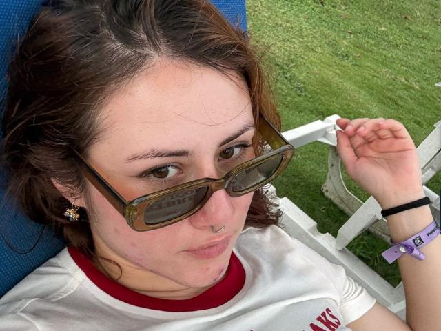 Close-up photo of MalenaJones lying down outdoors. She has wavy brown hair, light skin, and is wearing brown-tinted sunglasses, a white T-shirt with red trim, and small flower earrings. She has a nose ring and a purple wristband on her right wrist. She looks directly at the camera with a slightly neutral expression. The background shows green grass and a white fence. The blue chair she's lying on is partially visible.