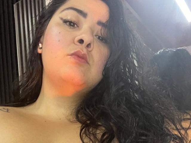 BoraLajuana with wavy black hair, light skin, and a slight red blush on her cheek, looking up with a neutral expression, small tattoo on her left shoulder.
