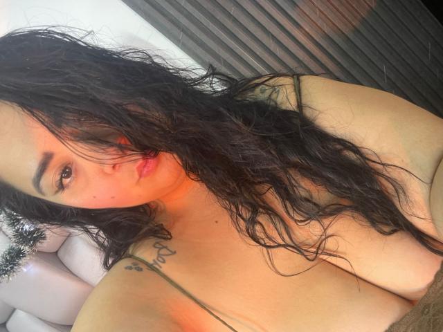 BoraLajuana with wet black hair, looking at the camera, topless, showing large tits, tattoos on her shoulder, and a slight pout, in a softly lit room.