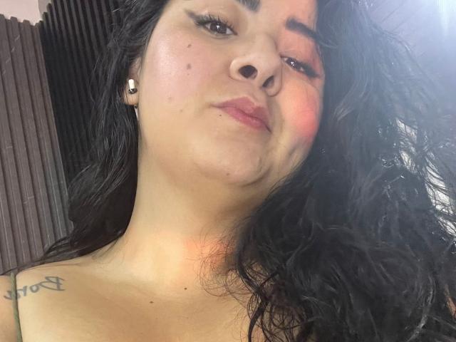 BoraLajuana's face, showing dark hair, black nose ring, tattoo on right shoulder, subtle makeup, and a slight pout, with pinkish cheek blush and a confident, relaxed expression.