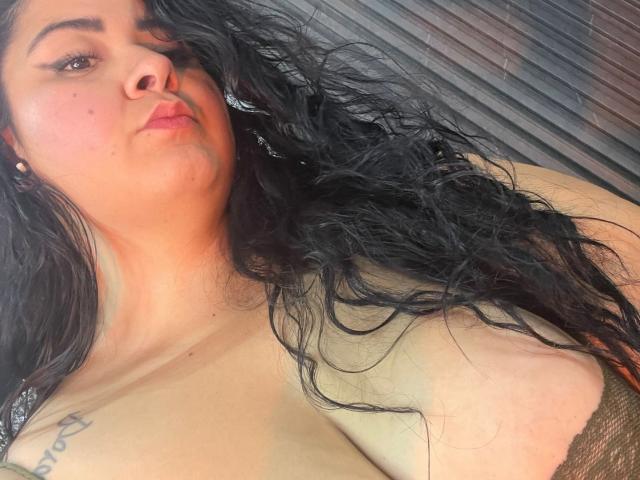 BoraLajuana with wavy black hair, light brown skin, and pouty lips, showing her bare tits and tattoo, wearing a brown lace bra, looking at the camera.