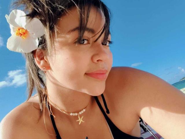 Close-up of KimDevinne with light brown skin, dark brown hair, and a white plumeria flower in her hair. She wears a black bikini top, a pearl choker, and a starfish necklace. Her dark hair is slightly messy, with bangs covering her forehead. She has a small smile and is looking to the side. Bright blue sky in the background. KimDevinne's facial features include a small nose and full lips. The image is a sunny, beach-style photograph.