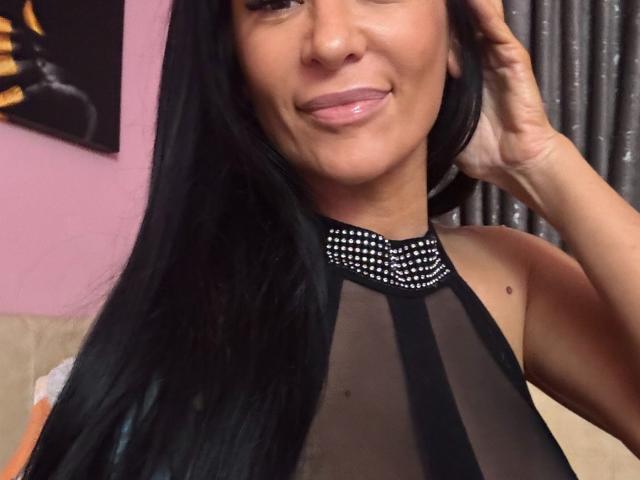 AndraD with long black hair, smiling, wearing a sheer black top with silver studs, hand in hair, showing her shiny pink lips and small mole on her right cheek.