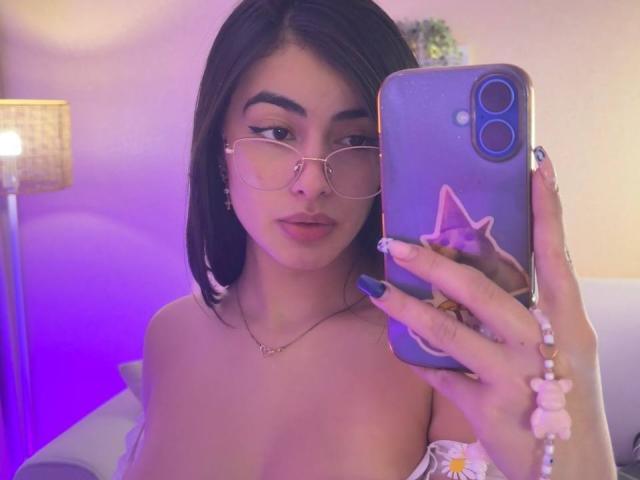 EmmaBeaumont takes a selfie, holding a phone with a purple case featuring a cat face sticker. She has long dark hair, wears glasses with thin gold frames, and has full pink lips. Her nails are long and painted blue. She's topless, showing her shoulders and a necklace with a small heart pendant. The room has a purple and pink light, a lamp on the left, and a potted plant in the background. Her expression is neutral.