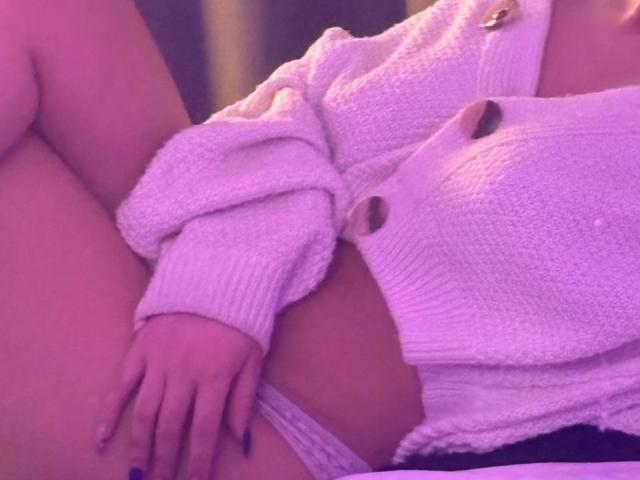 EmmaBeaumont with purple-pink tinted skin, wearing a white knitted cardigan, exposing her pussy with white lace panties, sitting with legs up, hand on her thigh.