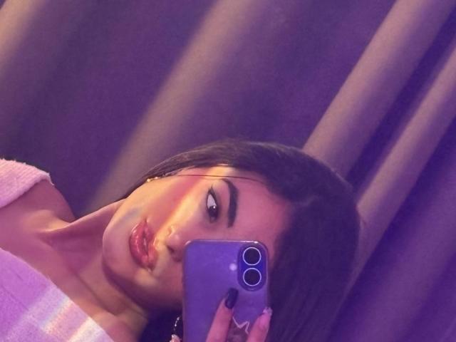 EmmaBeaumont takes a selfie with a purple iPhone, her face partially shadowed by the phone. She has dark, straight hair and pronounced eyebrows. Her lips are glossy and slightly puckered. She wears a white, off-the-shoulder top. Her nails are painted black. The light casts a warm, purple hue over the image. The focus is on her face and the phone. Her expression is neutral. The texture of the curtain in the background is smooth.