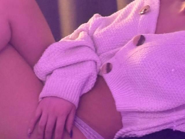 EmmaBeaumont's pink sweater covers her tits, showing a bit of her ass and panties, with her hand resting on her thigh, under purple lighting.