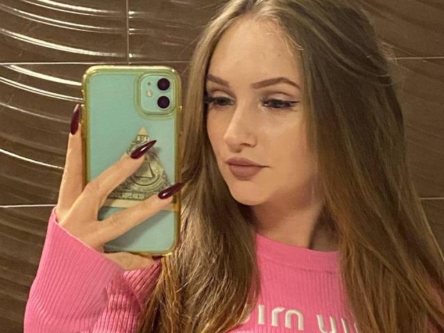 PamelaXu with long brown hair, wearing a pink sweater, holding a green phone with gold trim, nail polish is dark red, looking at herself in the mirror.