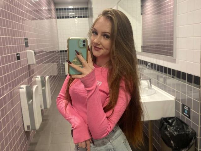 PamelaXu, a young woman with long, straight, light brown hair, stands in a pink-tiled bathroom, taking a mirror selfie. She wears a pink rib-knit sweater with a small logo on the left sleeve and light blue jeans. She holds a green phone with a pink case in her right hand. Her nails are dark red. The bathroom has white sinks, black tile accents, and a black trash can. Her smile is subtle. The image is a selfie with clear focus on her.