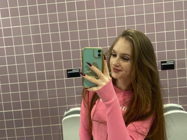 PamelaXu with long brown hair, pink sweater, and red nails, taking a selfie in a pink-tiled bathroom, holding a green phone, smiling slightly.