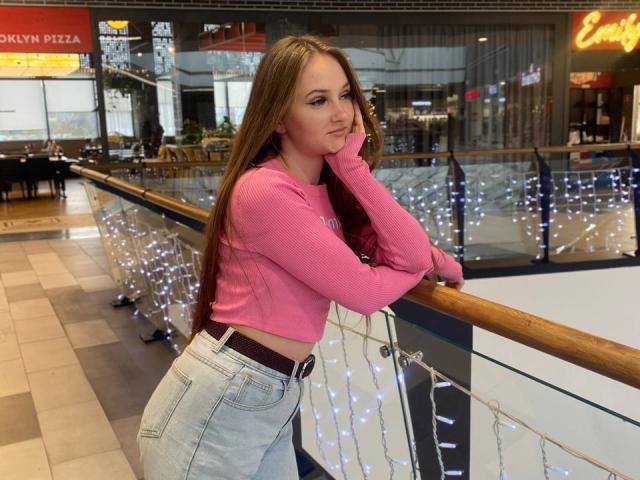 PamelaXu with long brown hair, pink ribbed crop top, and high-waisted jeans, leaning on a railing with white string lights, looking slightly annoyed, hand on cheek.
