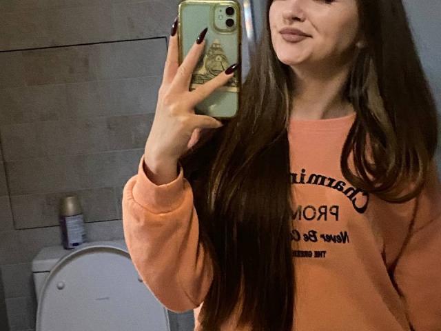 PamelaXu with long brown hair, wearing an orange sweatshirt, taking a mirror selfie with black nails, holding a green phone, showing a pouty face in a bathroom.