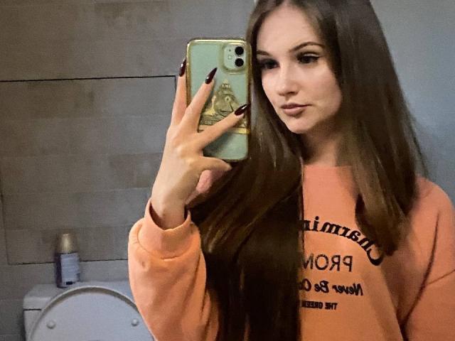 Pamela Xu, con cabello castaño largo, vistiendo una sudadera naranja, tomando una selfie en el espejo con un teléfono verde, uñas negras y una ligera mueca. Un inodoro se ve en el fondo..