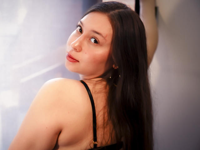 WidnyBeaumont with long brown hair, looking over her shoulder, wearing a black strap top, showing her bare back and small nose ring, with a slight smile.