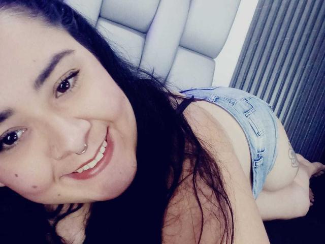 BoraLajuana with long black hair, smiling nose ring, wearing denim shorts, showing her ass, kneeling on black surface, blue eyes, light skin, small tattoos, casual selfie.